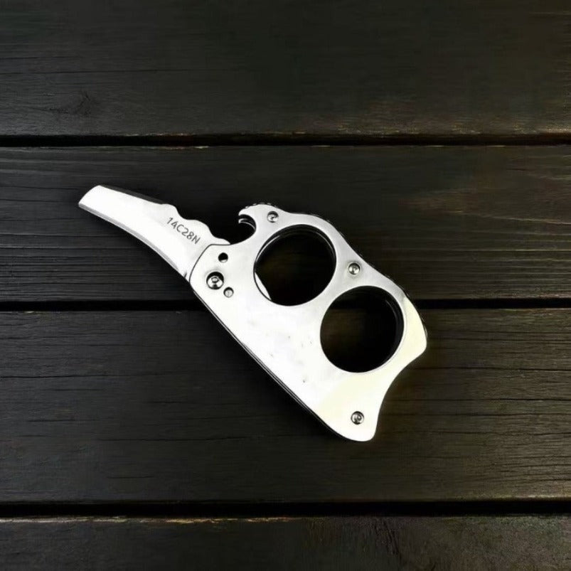 Stainless steel mini knuckle knife with hawkbill blade resting on dark wood surface